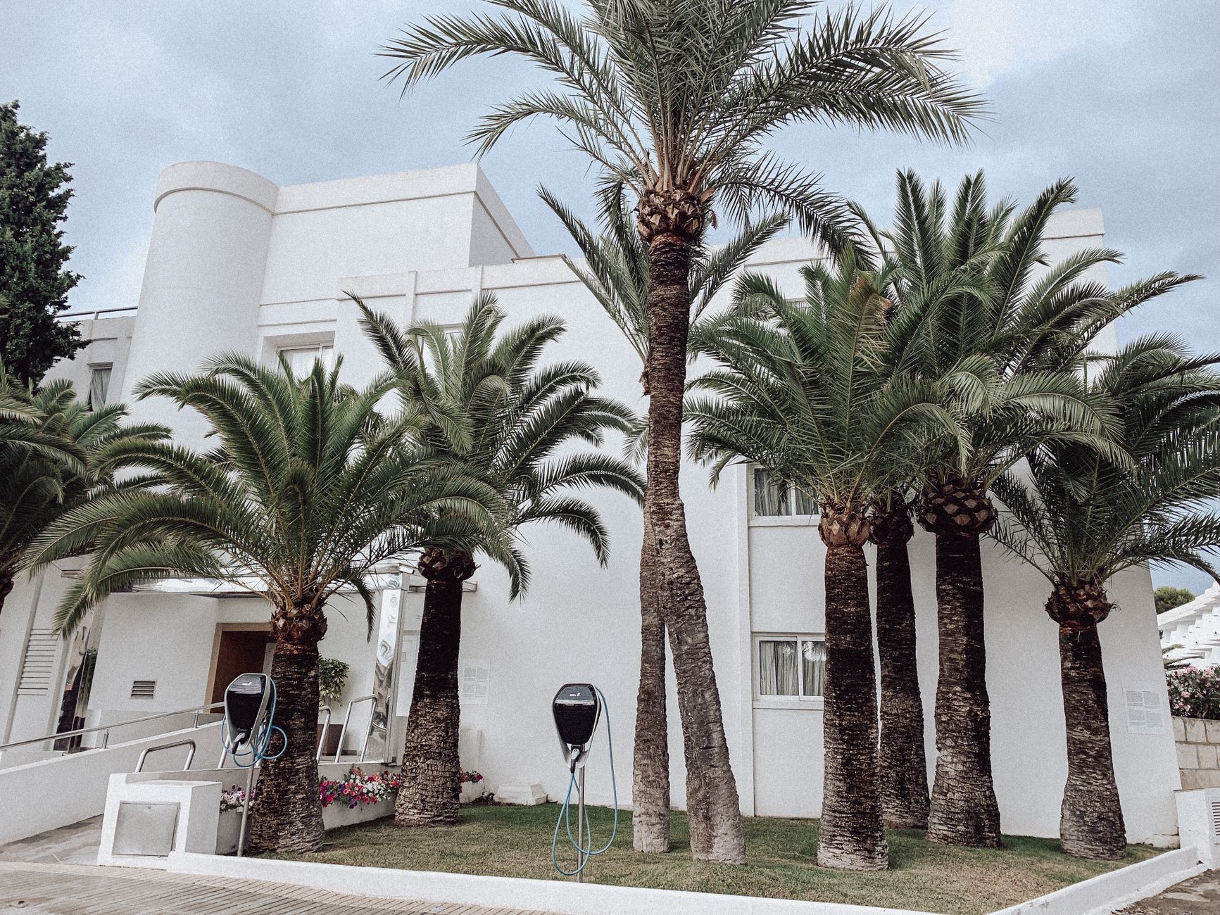 Contemporary white building facade with palm trees and charging stations on a city street.