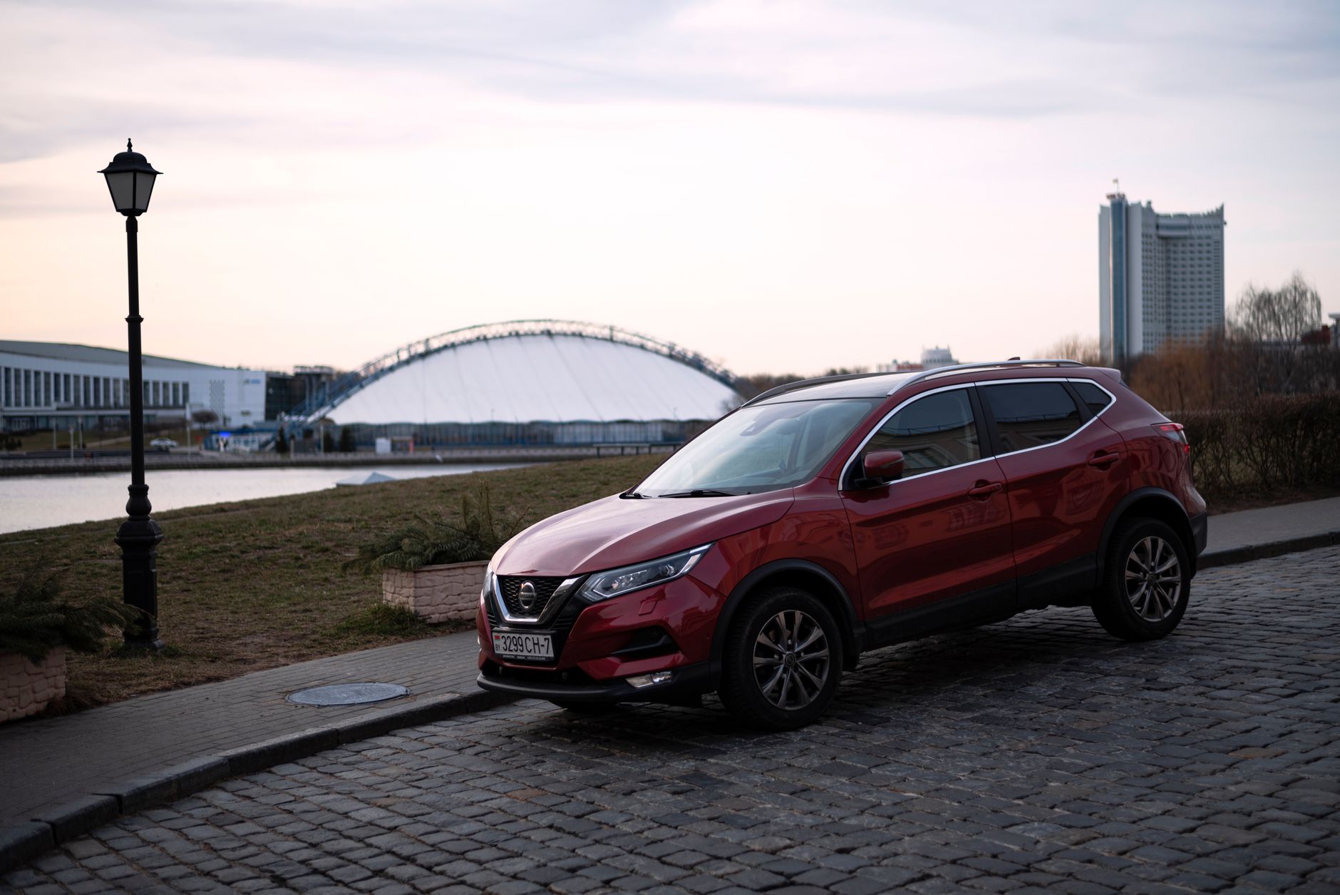A red Nissan Qashqai parked on cobblestone streets near the river in Minsk cityscape.