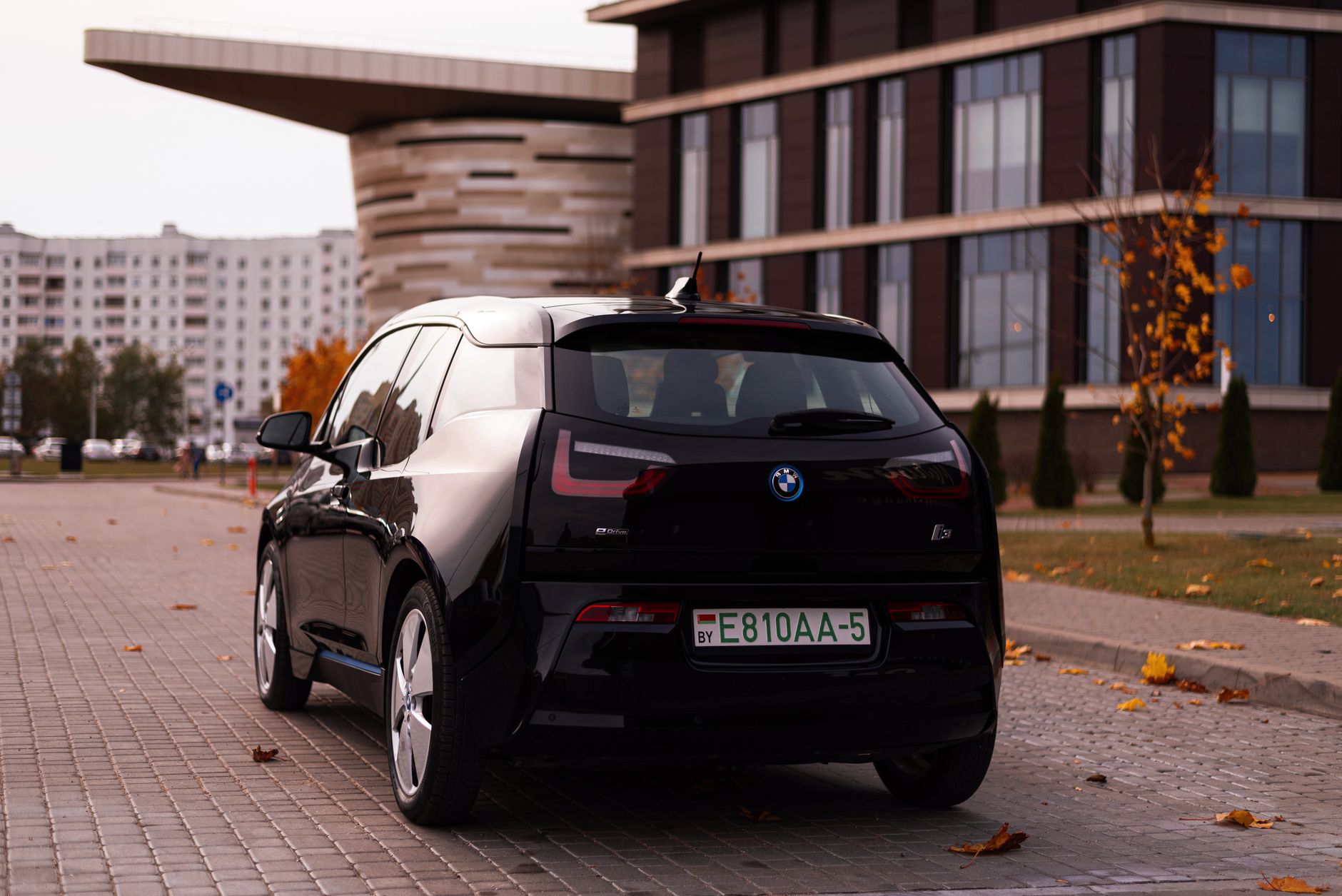 Sleek BMW i3 car parked on a city street with autumn leaves and modern buildings.