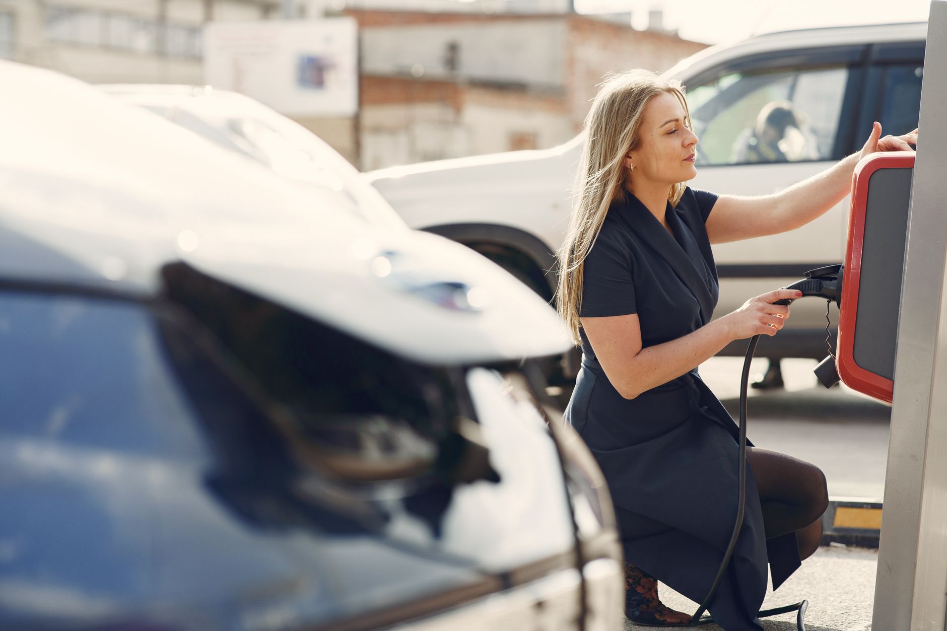 Side view of full length female driver in casual dress squatting near station while charging electric automobile for continuation of driving