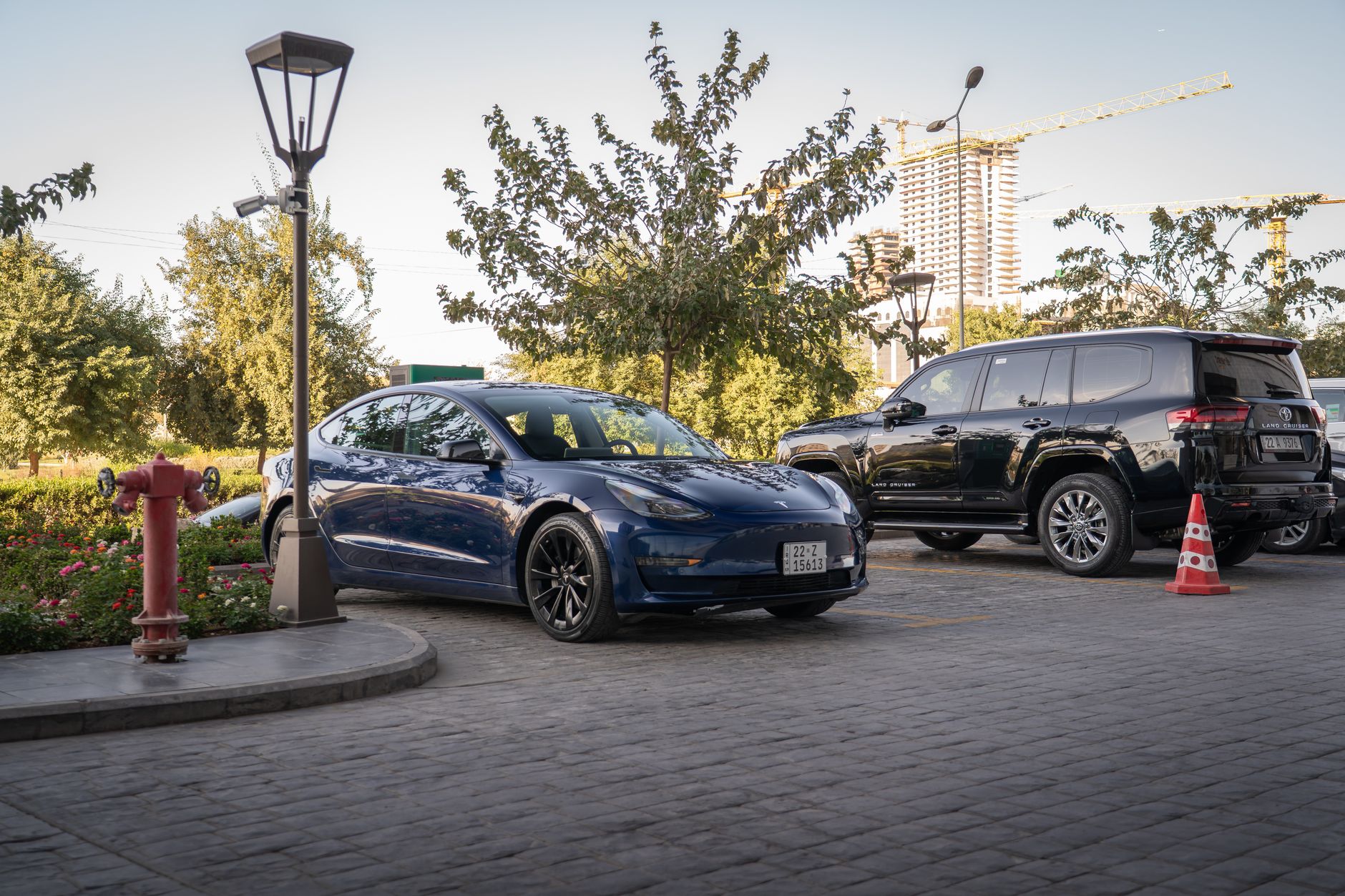 A sleek Tesla Model 3 electric car parked in a sunny urban parking lot with greenery and streetlights.