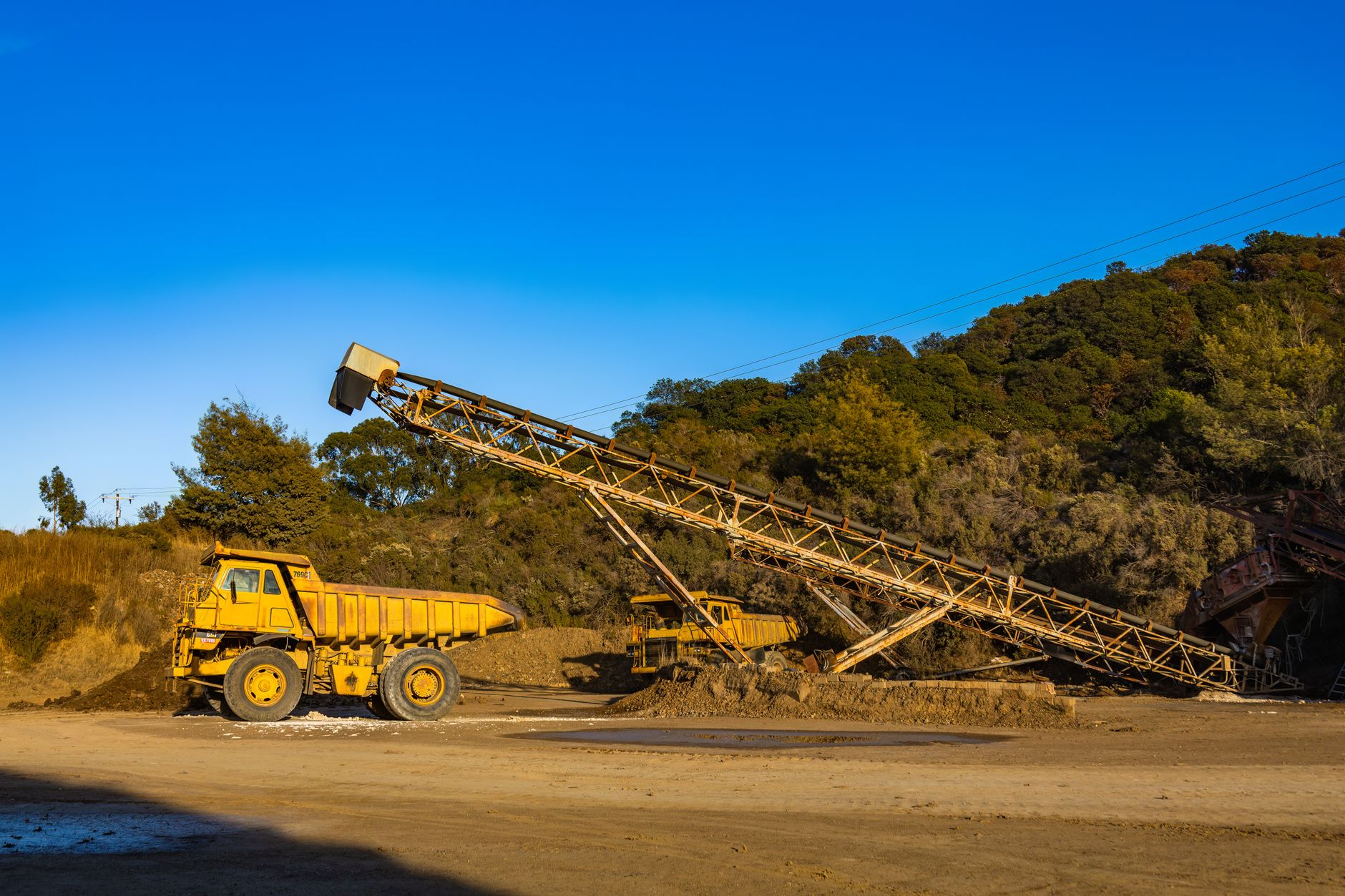Yellow dump trucks and conveyor machinery in a sunny mining site with clear blue sky.
