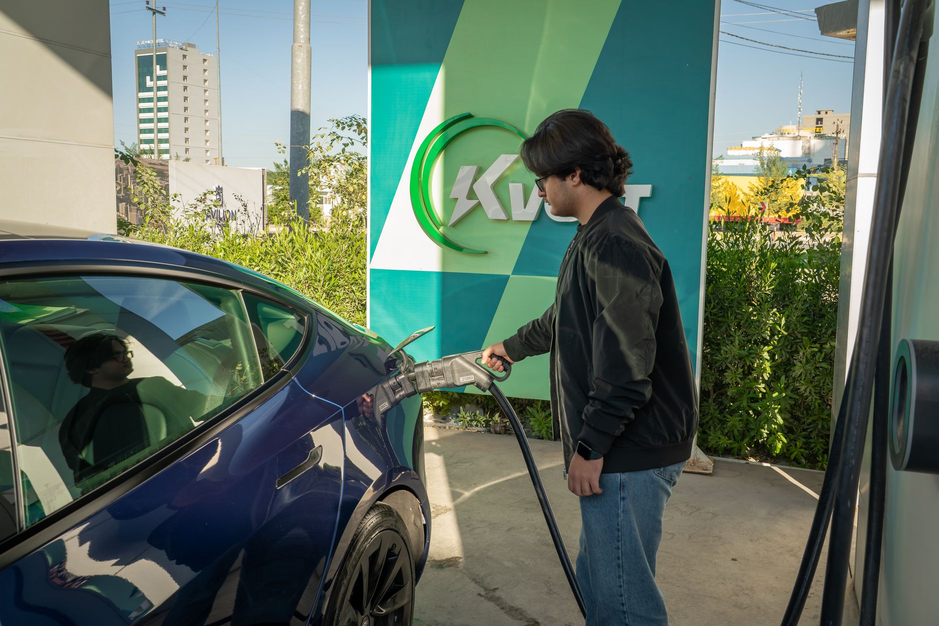 Young man charging a Tesla Model 3 at an outdoor electric vehicle station during the day.