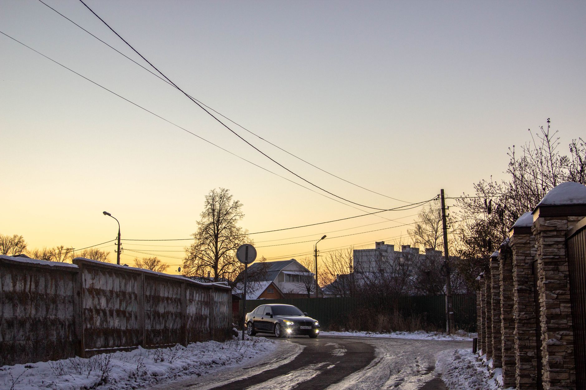 A sleek car drives on a snow-covered city road during a winter sunset, creating a serene ambiance.